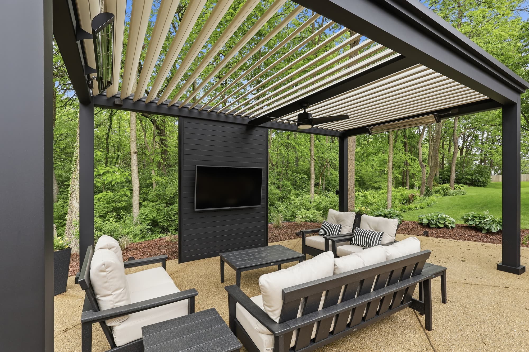 A louvered pergola with cream-coloured louvers, mounted overhead heaters, a ceiling fan and a wall-mounted television above an outdoor lounge seating area.