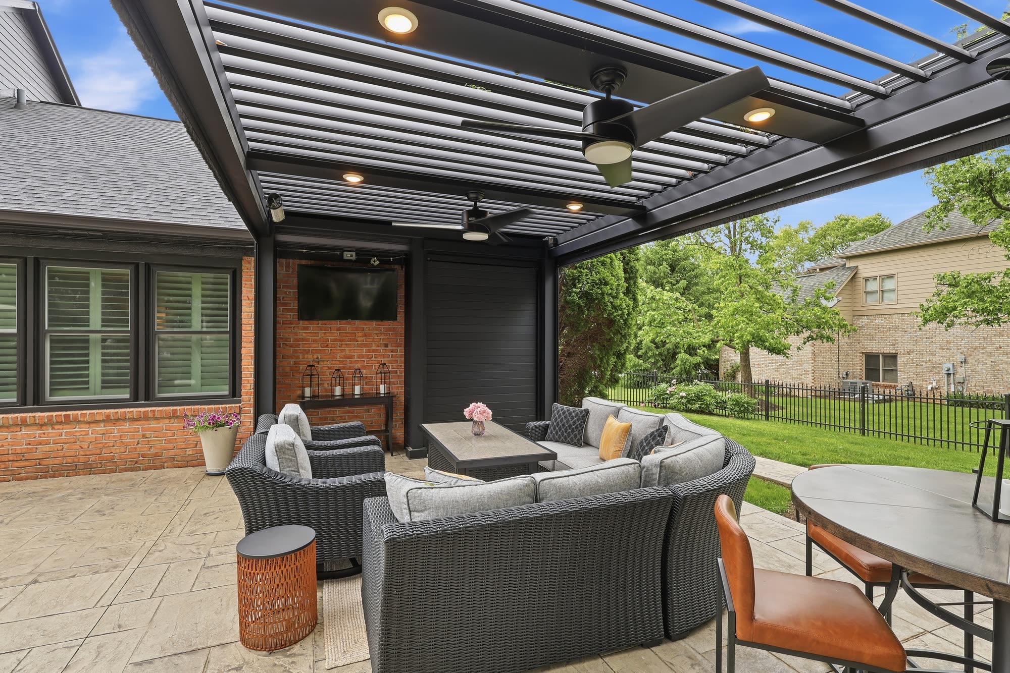 Open louvers on a pergola with ceiling fans and recessed lighting above a curved outdoor sofa and dining set on a stamped concrete patio.